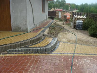 Układanie kostki brukowej w dwóch kolorach (czerwony i żółty) z elementami schodów, widoczny mikser do betonu i wąż ogrodowy.